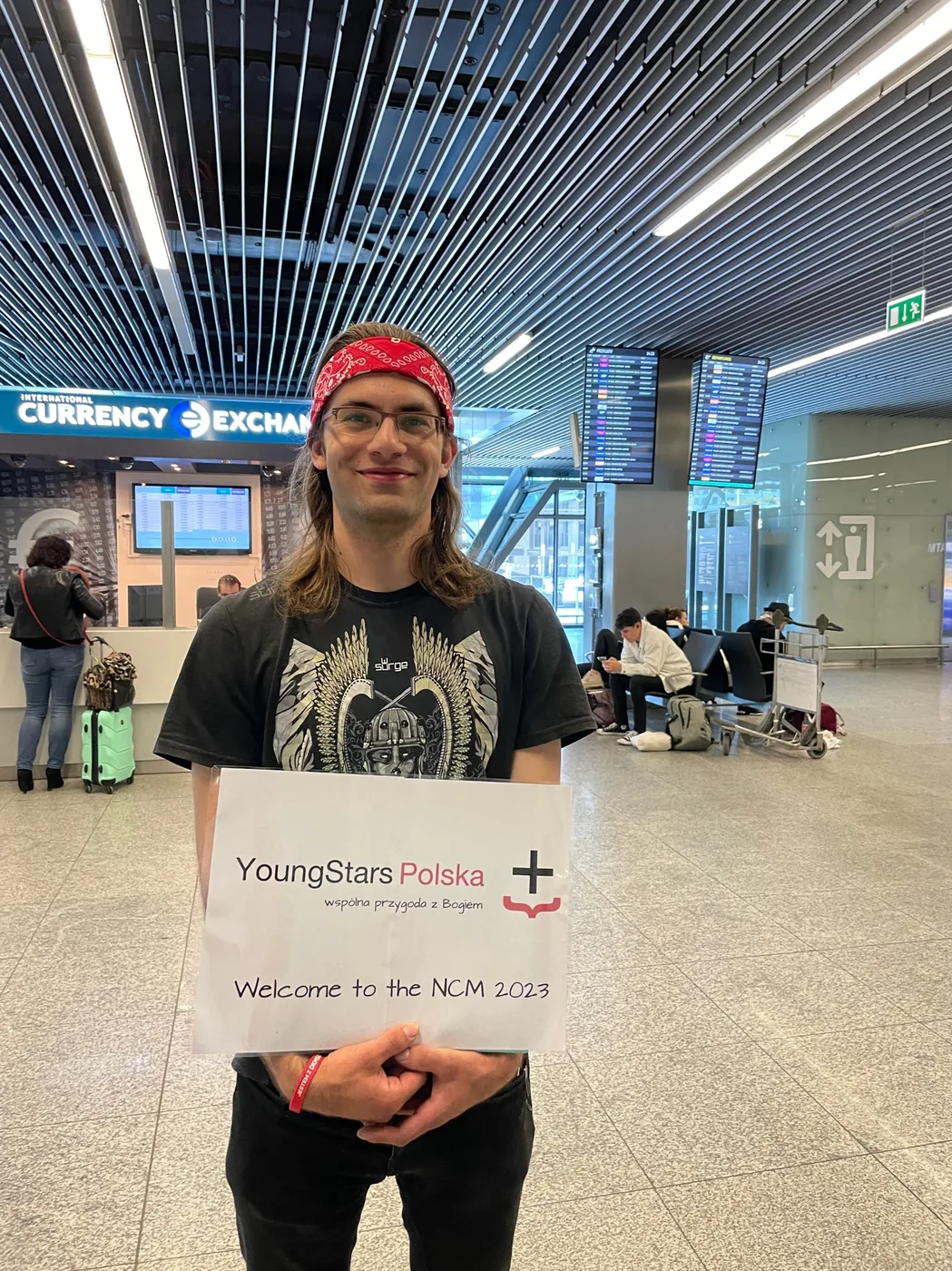 Junger Mann mit Schild «YoungStars Polska, Welcome to the NCM 2023» am Flughafen
