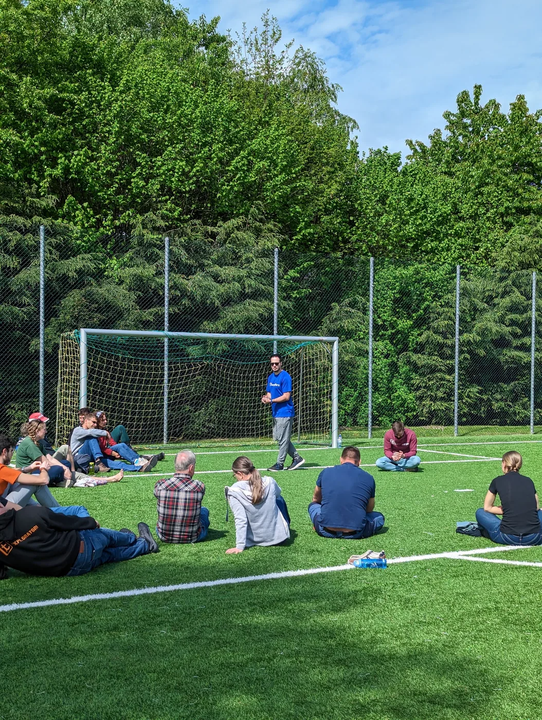 Ein Leiter spricht zum sitzenden Kreis auf dem Sportplatz, alle hören aufmerksam zu