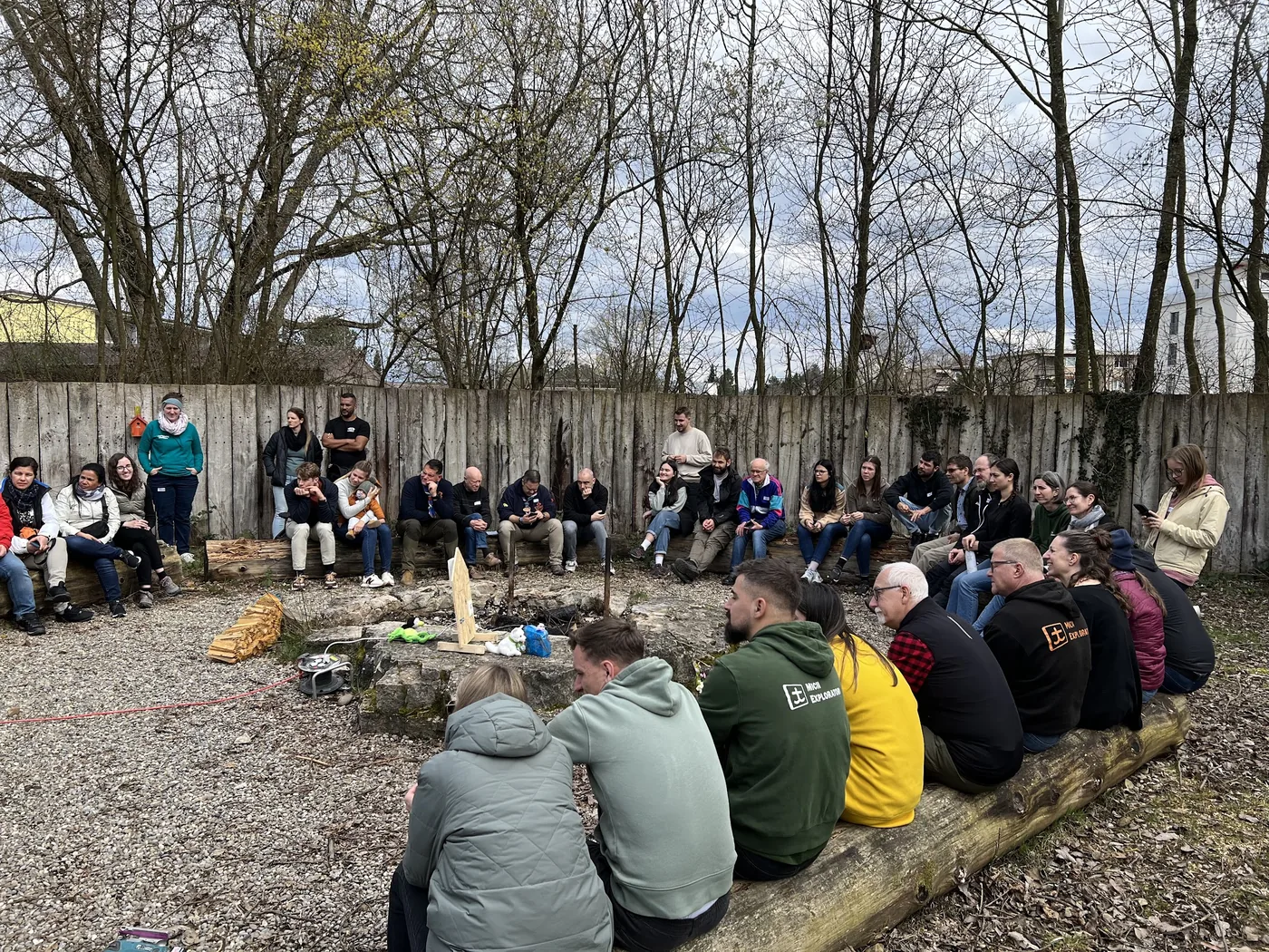 Grosse Gruppe sitzt im Kreis auf Baumstämmen um eine Feuerstelle im Wald