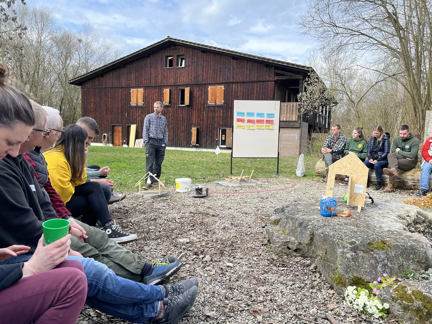 Outdoor-Workshop vor dem Holzhaus: Leiter erklärt Farbkarten-Tafel, Gruppe hört im Kreis sitzend zu