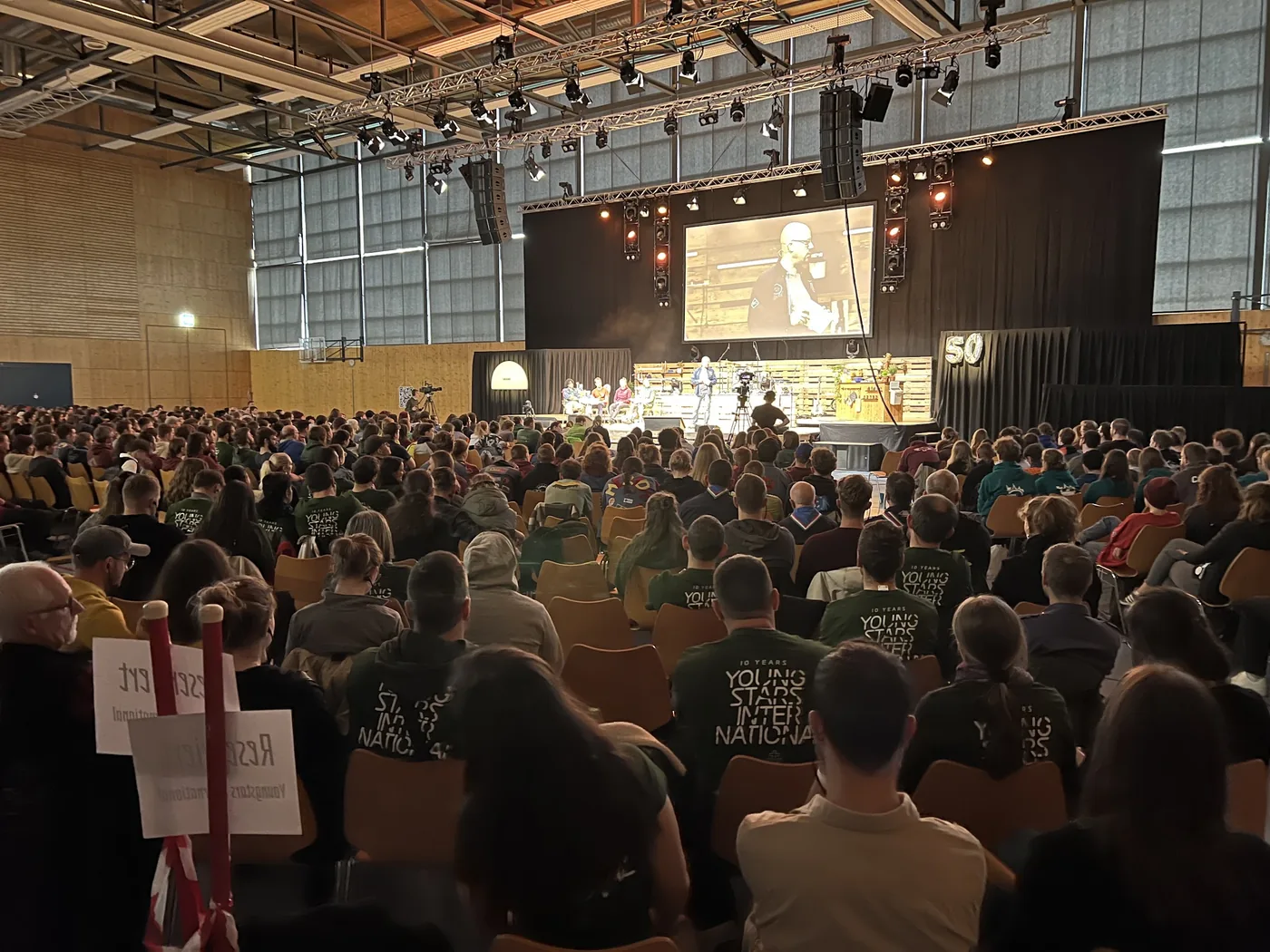 Grosse Abendveranstaltung in der Halle: volles Publikum mit Youngstars-Hoodies vor Bühne und Leinwand