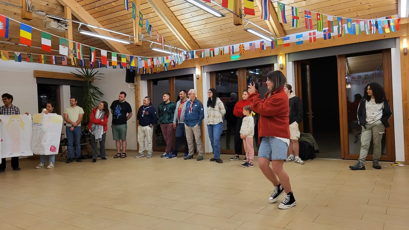 Frau in roter Jacke in Bewegung vor der Gruppe im flaggengeschmückten Saal, alle schauen zu