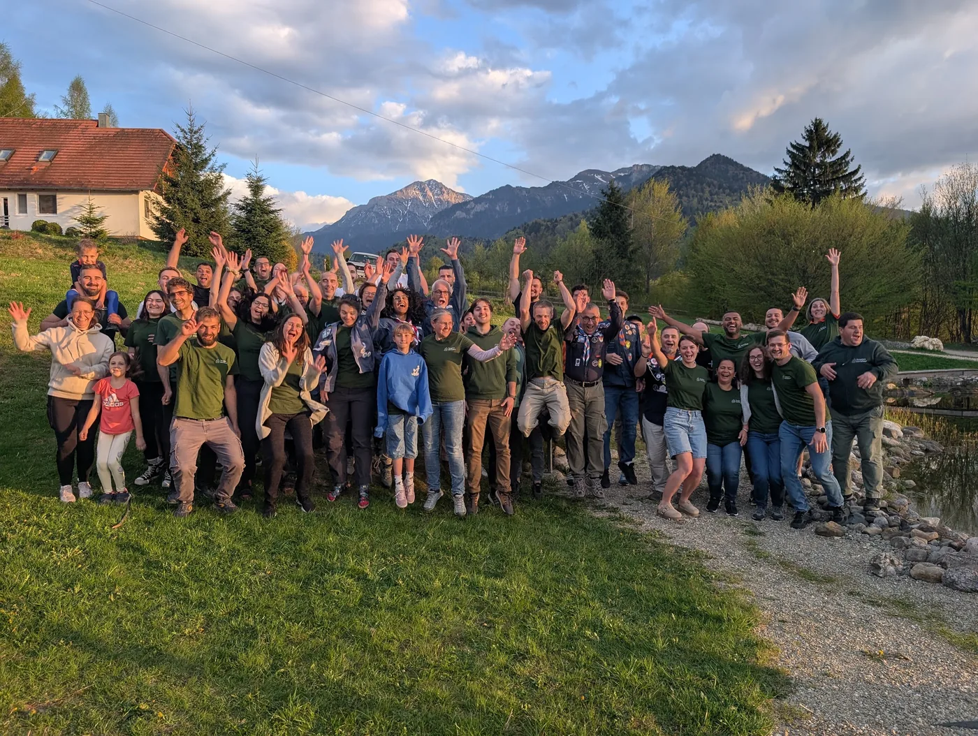 Grosses Gruppenfoto der gesamten Konferenz am Teich, jubelnd mit erhobenen Händen, Berge und Abendlicht