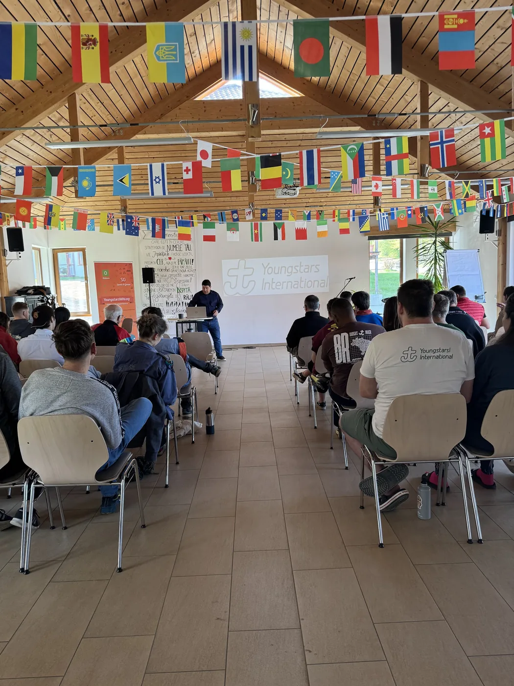 Plenumsaal von hinten: Teilnehmende unter dutzenden Nationalflaggen, Youngstars-International-Projektion vorn