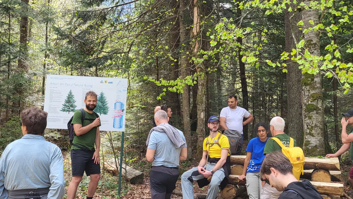 Gruppe hört einem Führer an der Infotafel des Naturparks Bucegi im Wald zu, Teilnehmende sitzen auf Baumstämmen
