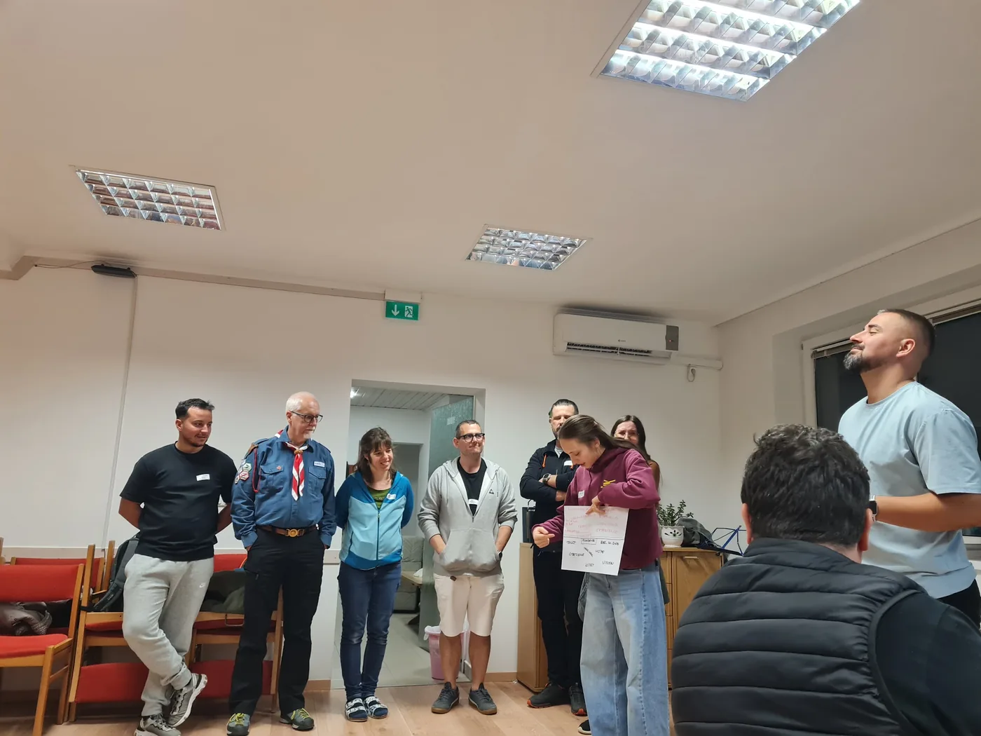 Internationale Gruppe im Seminarraum, eine Teilnehmerin erklärt ihr Plakat, Pfadfinder-Kluft eines Partnerlands sichtbar