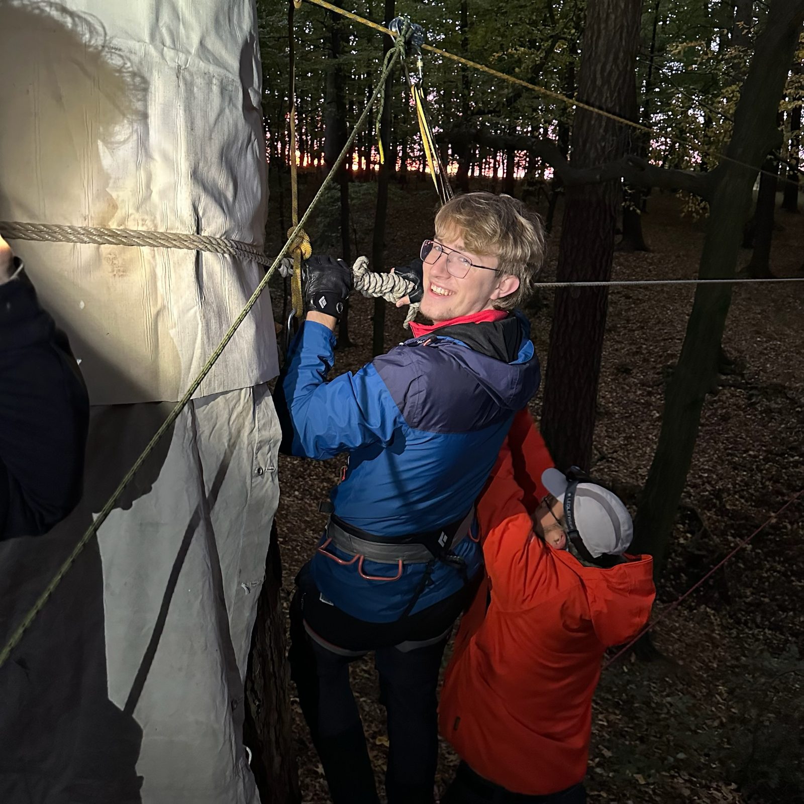Ein Teilnehmer überquert lachend eine selbst gebaute Seilbrücke im Wald.